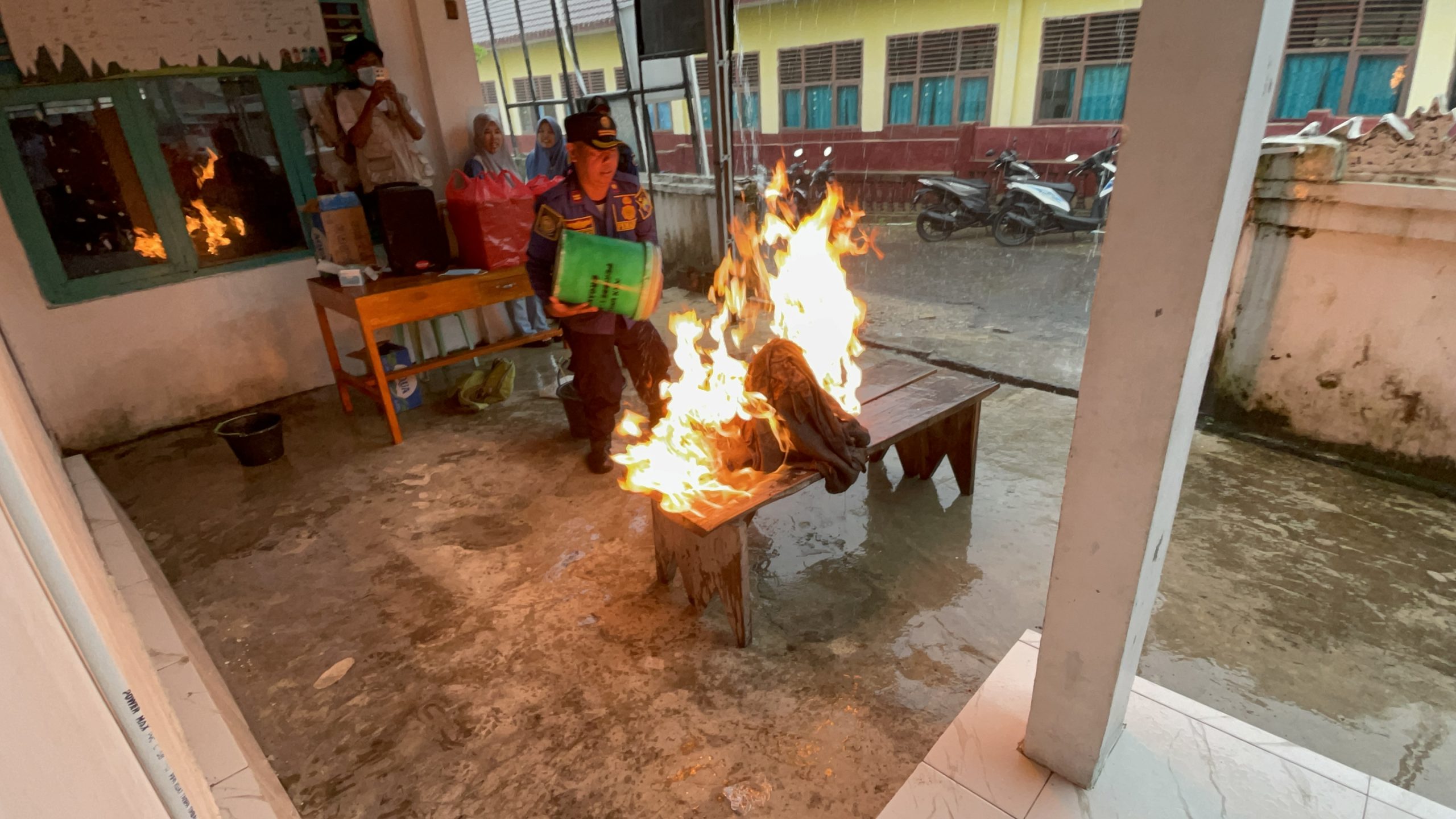 Salah satu tim pemadam kebarakaran sedang memperagakan cara memadamkan api pada kebakaran kecil di rumah. Beberapa orang berdiri di belakangnya menyaksikan dan memotret