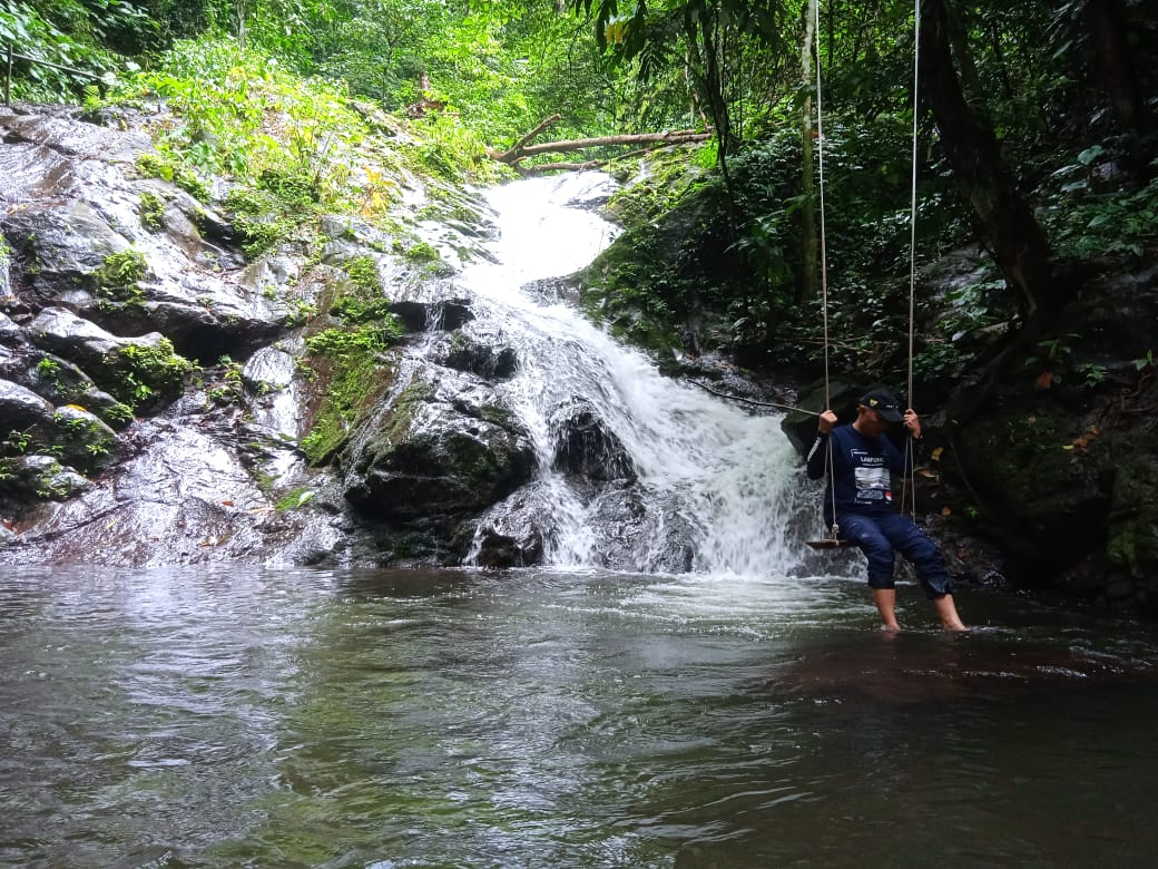Seorang laki-laki sedang duduk di ayunan dengan latar pemandangan air terjun 9 Puteri