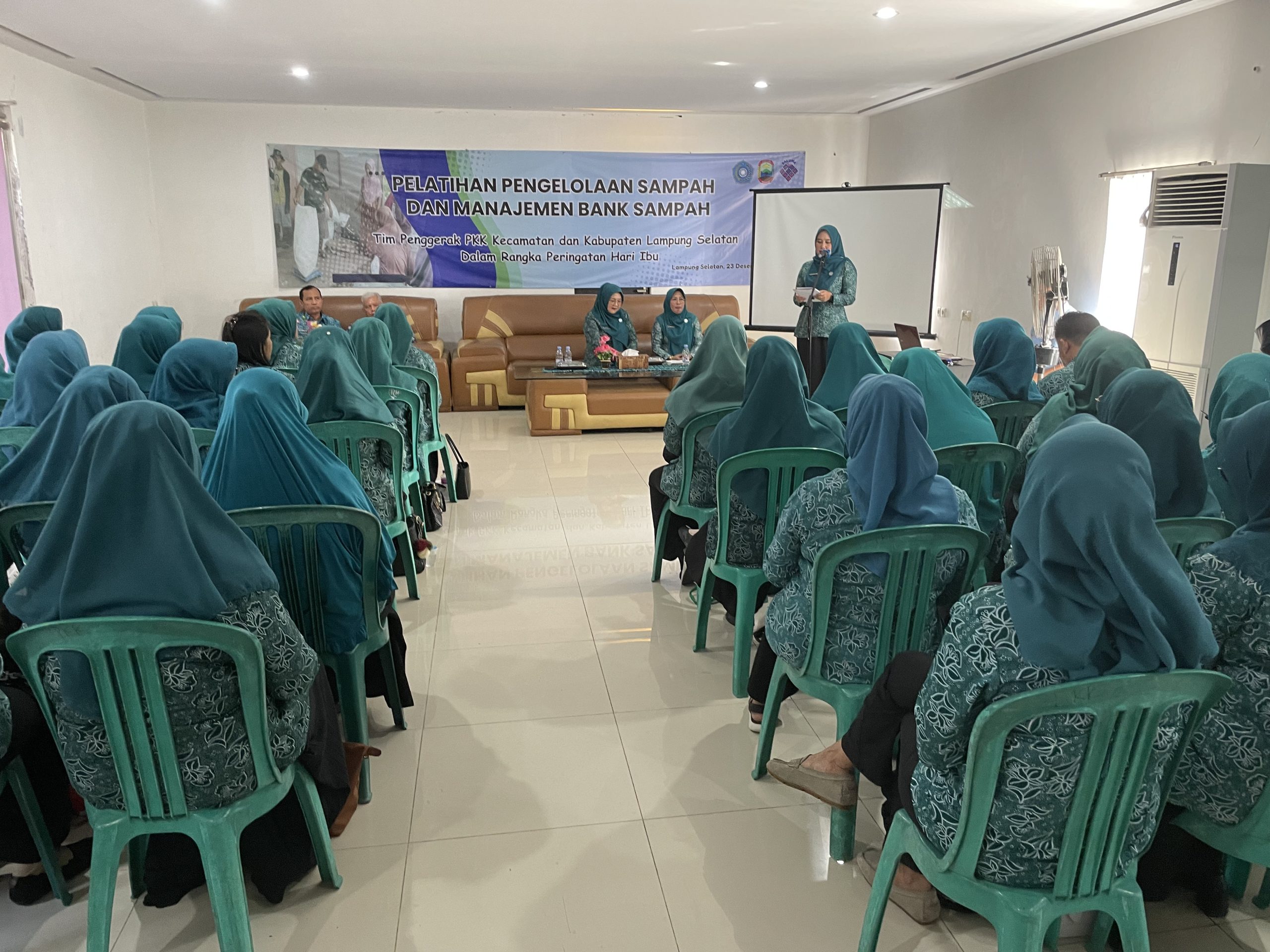 Ibu-ibu sedang duduk di ruangan menyimak satu ibu yang sedang berdiri di depan mic