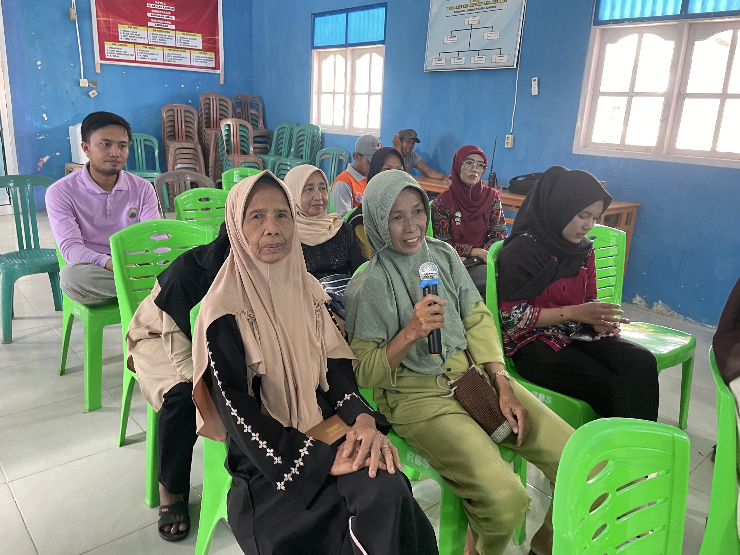 Sejumlah peserta perempuan dan laki-laki duduk di kursi plastik hijau di dalam aula desa berdinding biru, mengikuti kegiatan lokakarya; seorang perempuan berhijab hijau memegang mikrofon dan menyampaikan pendapat, sementara peserta lain mendengarkan dengan penuh perhatian.