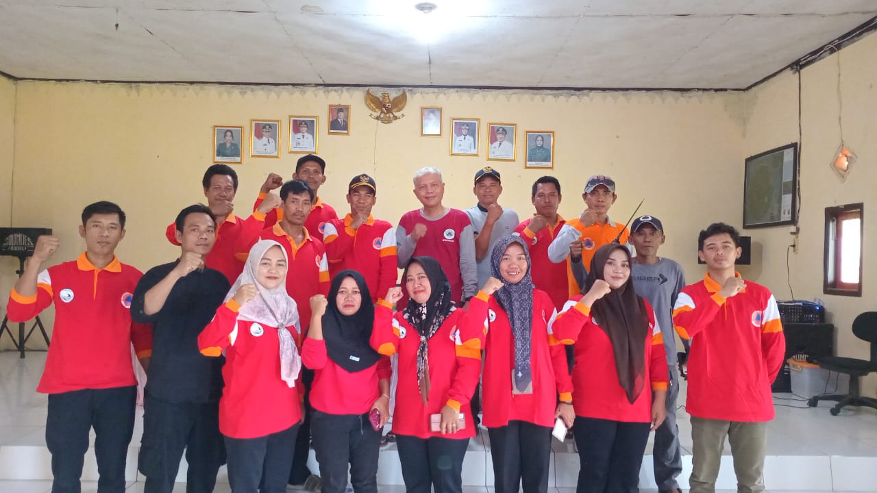 Foto bersama sekelompok relawan dan aparat desa mengenakan kaos merah berdiri di dalam aula balai desa, berpose mengangkat tangan mengepal sebagai simbol semangat dan solidaritas, dengan latar dinding berisi foto pejabat pemerintahan dan lambang Garuda.