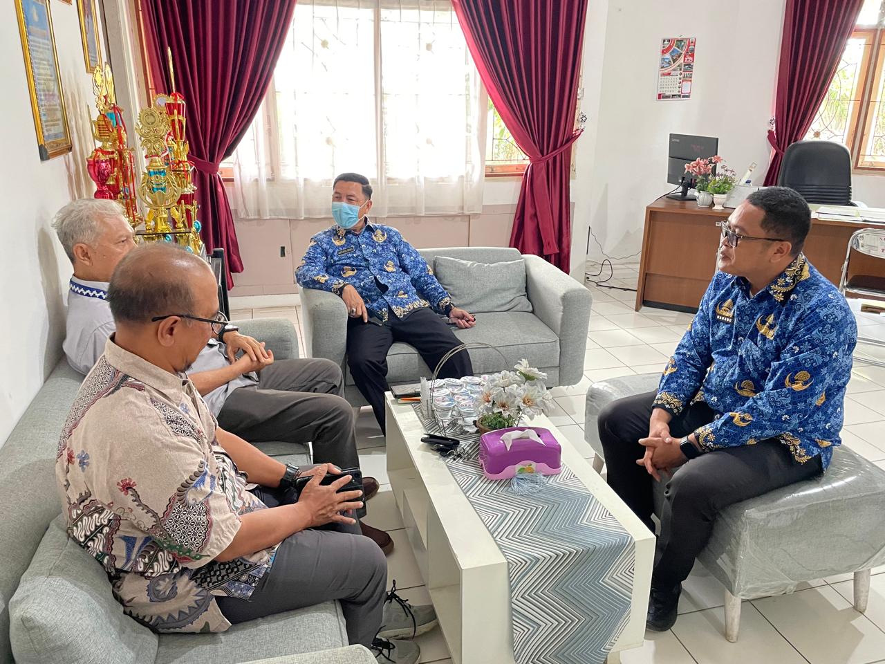 Empat pria mengenakan batik duduk berbincang di ruang kantor yang terang, dengan sofa abu-abu mengelilingi meja kecil berisi minuman dan tisu; di latar belakang terlihat rak piala, meja kerja, dan jendela dengan tirai merah marun.