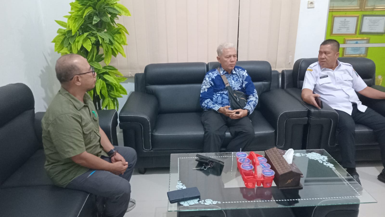 Tiga pria duduk berdiskusi di ruang kantor dengan sofa hitam dan meja kaca di tengah; dua mengenakan seragam putih dan satu mengenakan batik biru, dengan tanaman hias, tirai vertikal, dan foto pejabat di dinding sebagai latar belakang.
