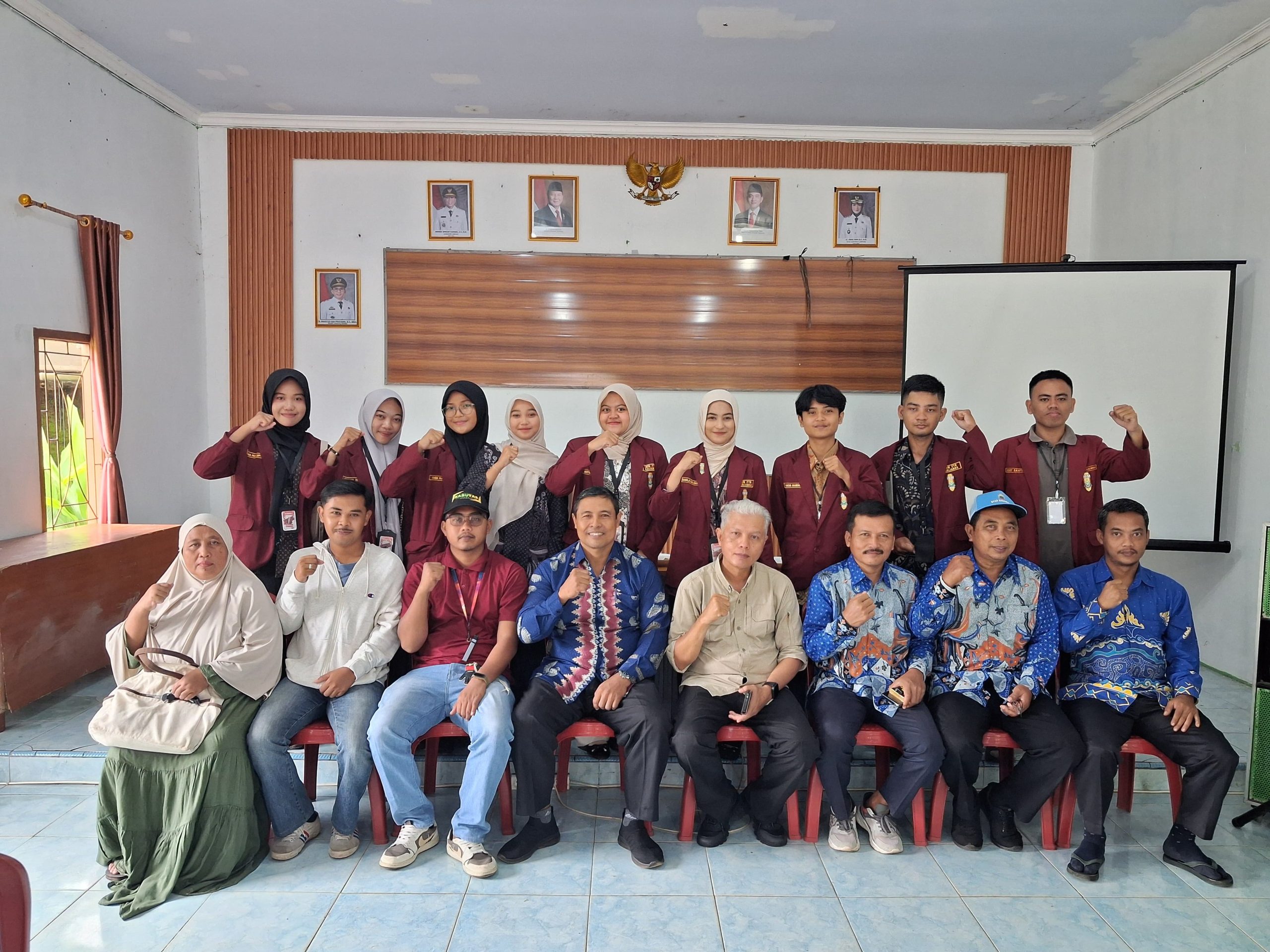 Foto bersama peserta Sosialisasi Pengelolaan Sampah di Aula Kantor Desa Sidomulyo. Sejumlah mahasiswa Ikatan Mahasiswa Muhammadiyah Lampung Selatan mengenakan jas almamater marun berdiri di barisan belakang, sementara warga desa, perangkat desa, dan narasumber duduk di barisan depan. Seluruh peserta mengangkat tangan mengepal sebagai simbol semangat dan komitmen bersama. Di latar belakang terlihat lambang Garuda Pancasila, foto pejabat pemerintahan, dan layar proyektor.