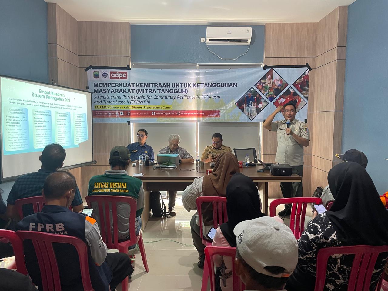 Suasana Workshop Sistem Komunikasi Peringatan Dini Tsunami Selat Sunda di aula Pos Pemantauan Gunung Anak Krakatau. Seorang narasumber dari BPBD Lampung Selatan berdiri menyampaikan materi di depan peserta, sementara tiga pemateri lainnya duduk di meja panel. Di layar proyektor terlihat presentasi tentang sistem peringatan dini berbasis komunitas untuk menghadapi ancaman tsunami non-tektonik Gunung Anak Krakatau. Peserta workshop terdiri dari perwakilan Destana dan warga pesisir yang duduk menyimak materi. Di belakang panel terpampang spanduk kegiatan SPRINT PALUMA Nusantara