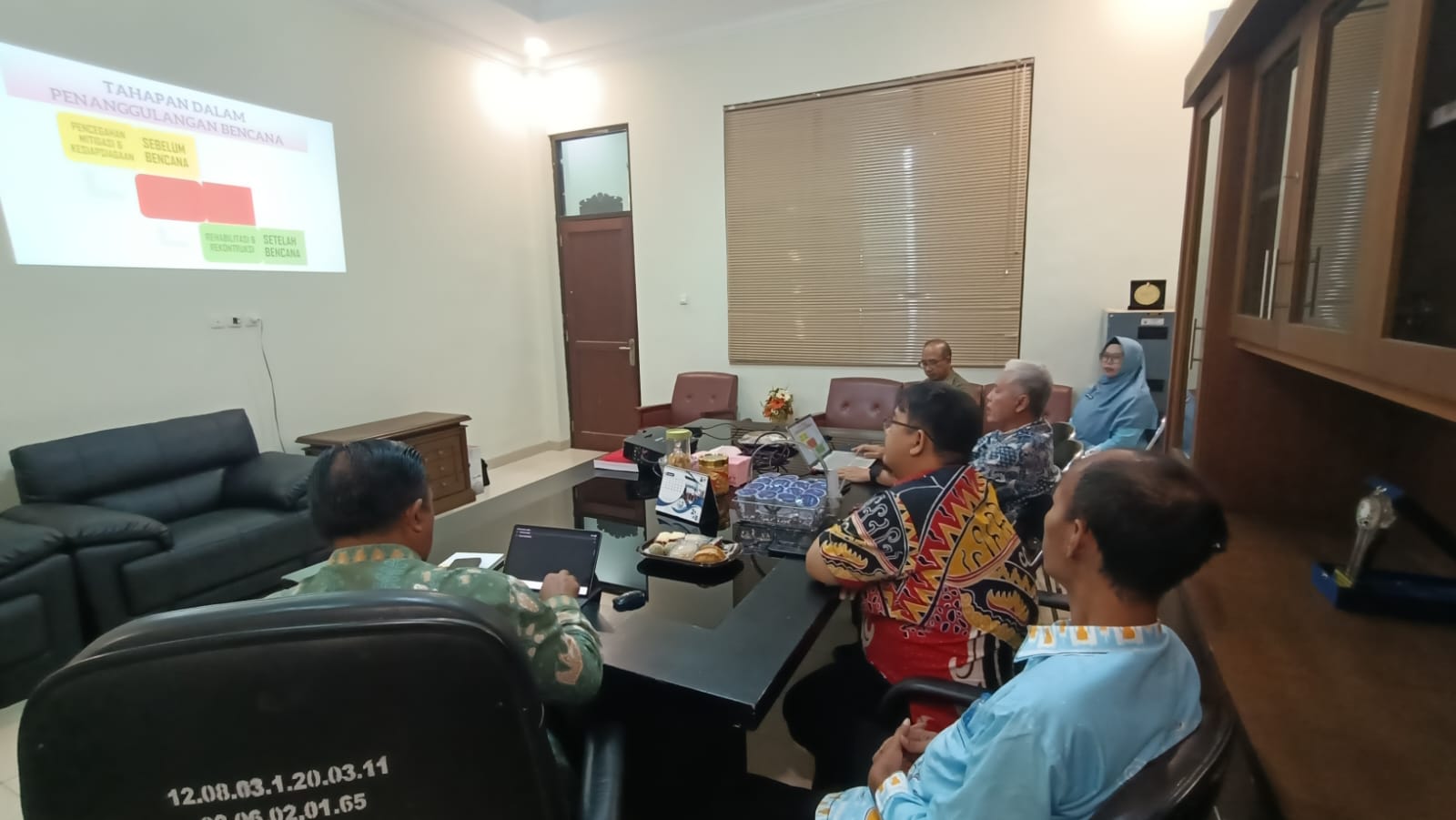 Sejumlah orang duduk mengelilingi meja rapat di sebuah ruang kantor, memperhatikan presentasi yang diproyeksikan di dinding bertuliskan “Tahapan dalam Penanggulangan Bencana.” Beberapa peserta membawa laptop dan catatan, suasana rapat terlihat formal dan fokus, dengan perabot kantor seperti sofa, lemari arsip, dan jendela tertutup tirai di latar belakang.