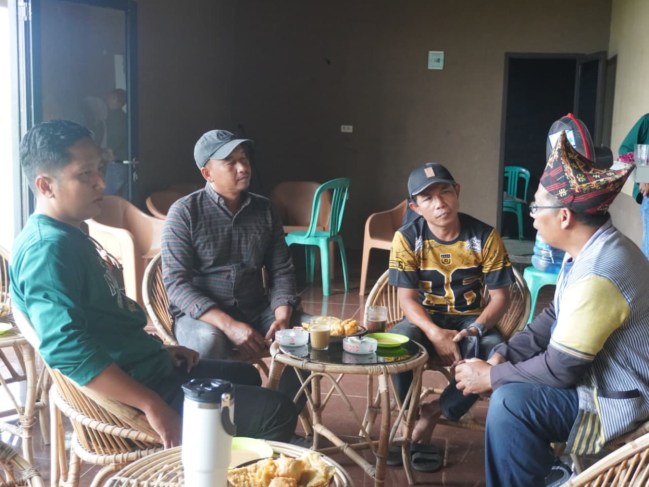 Empat pria duduk melingkar di kursi rotan sambil berdiskusi santai di sebuah ruangan semi-terbuka, dengan meja kecil berisi minuman dan camilan di tengah sebagai suasana pertemuan informal.
