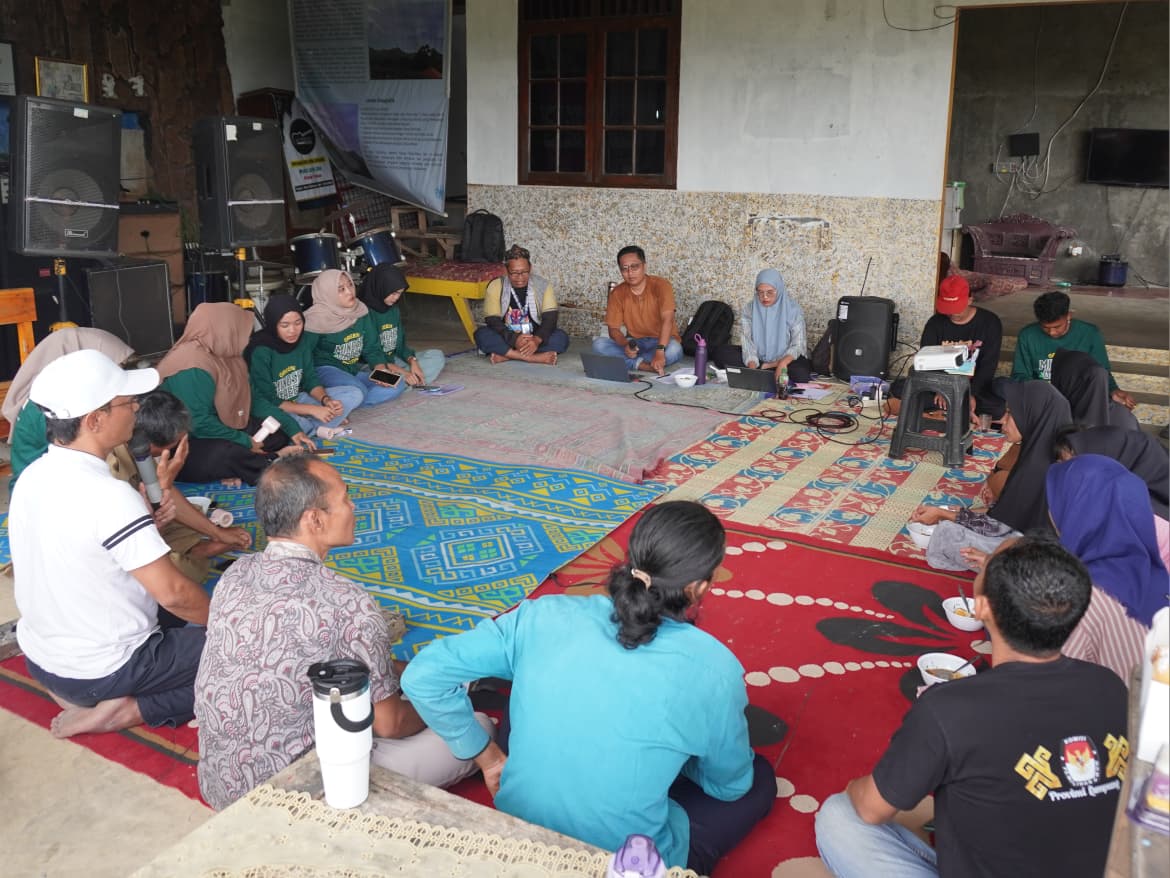 Para relawan dan fasilitator duduk melingkar di atas tikar dalam sebuah ruang terbuka, mengikuti diskusi atau lokakarya komunitas dengan perlengkapan presentasi dan pengeras suara di sekelilingnya