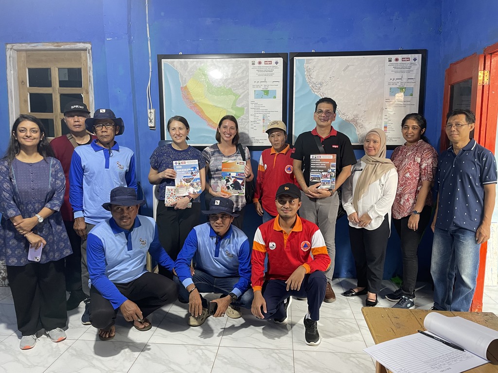 Sekelompok sekitar sebelas orang dewasa berfoto bersama di dalam ruangan bercat biru. Beberapa orang berdiri dan tiga orang berjongkok di bagian depan. Dua orang perempuan di tengah memegang buku atau laporan bergambar. Beberapa pria mengenakan seragam lapangan berwarna biru dan merah dengan logo instansi, sementara yang lain mengenakan pakaian kasual dan semi-formal. Di dinding belakang terdapat dua peta besar yang dipajang dalam bingkai. Di sisi kanan terlihat meja dengan buku tamu atau lembar absensi terbuka. Semua orang menghadap kamera dan tersenyum.