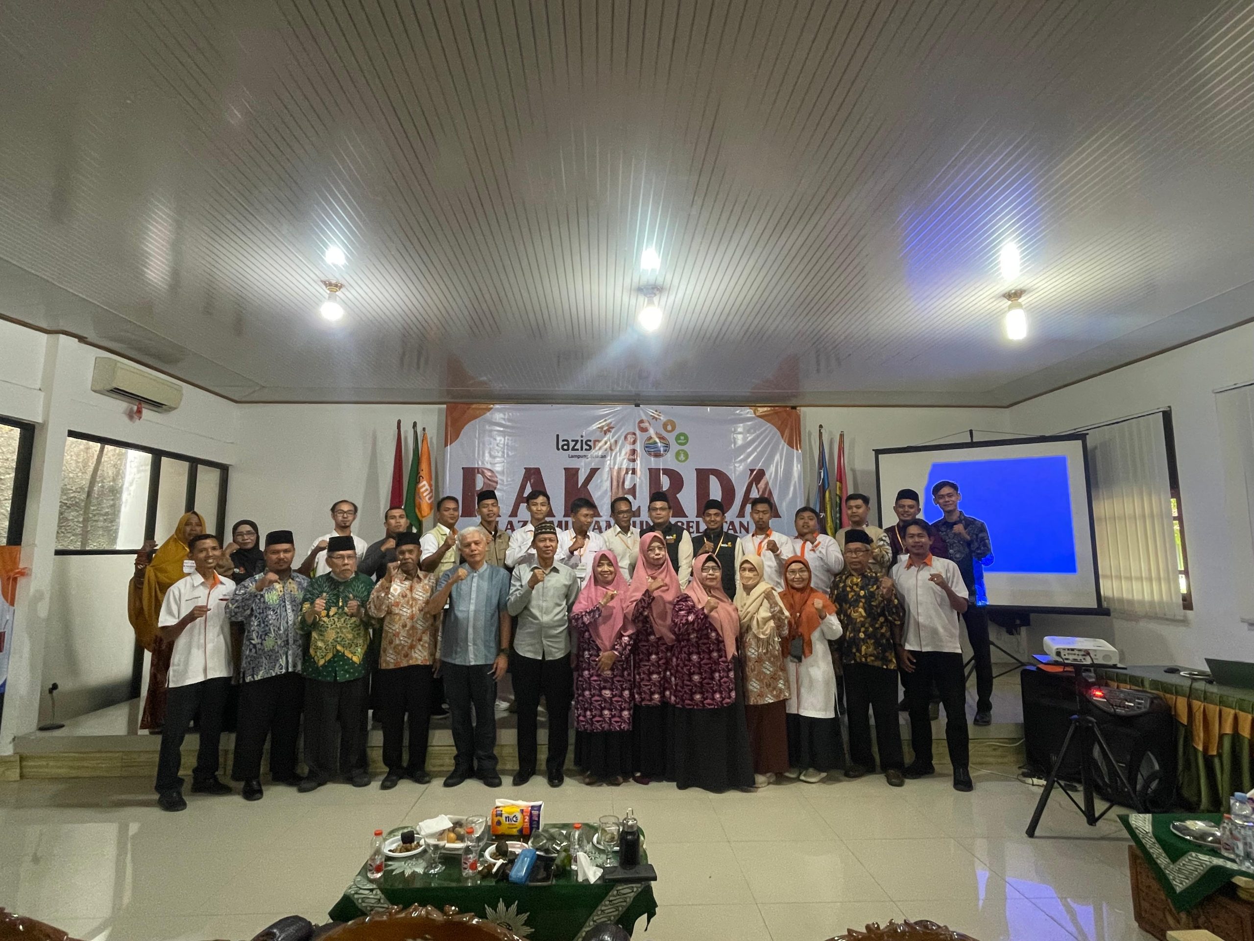 Foto bersama peserta dan pengurus dalam acara Rakerda LAZISMU Lampung Selatan di sebuah aula. Puluhan orang berdiri berjajar di depan backdrop bertuliskan “RAKERDA LAZISMU Lampung Selatan”, mengenakan beragam busana formal dan batik. Di belakang mereka terlihat bendera organisasi, layar proyektor, dan perlengkapan acara. Suasana tampak khidmat dan penuh kebersamaan, dengan peserta mengangkat tangan sebagai simbol semangat dan komitmen bersama.