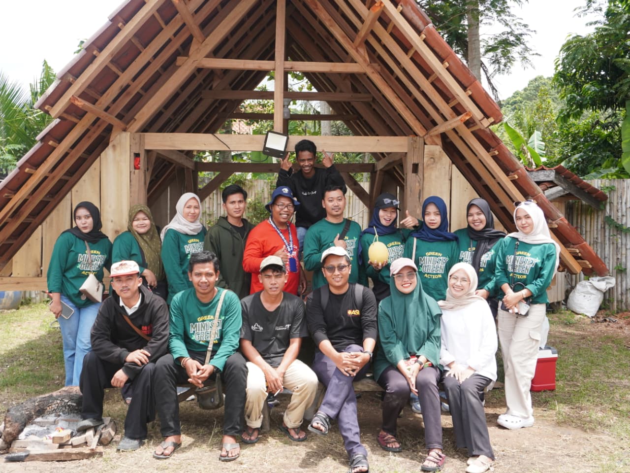 Sekelompok pemuda dan pendamping berpose bersama di depan bangunan kayu berbentuk saung di area kebun, mengenakan kaus hijau bertema lingkungan, dengan latar pepohonan dan suasana pedesaan.