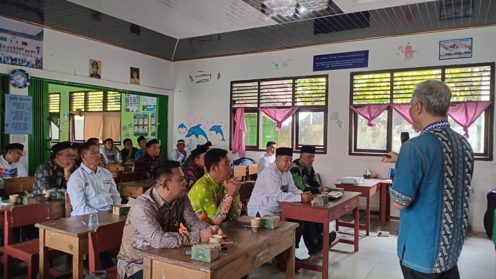 Seorang narasumber berdiri di depan kelas memberikan pemaparan kepada peserta yang duduk di bangku-bangku kayu seperti ruang kelas sekolah. Para peserta, sebagian mengenakan batik dan peci, terlihat menyimak dengan serius. Ruangan memiliki jendela besar dengan tirai pink, dinding dihiasi poster edukasi dan gambar, menciptakan suasana diskusi atau pelatihan komunitas yang formal namun sederhana.