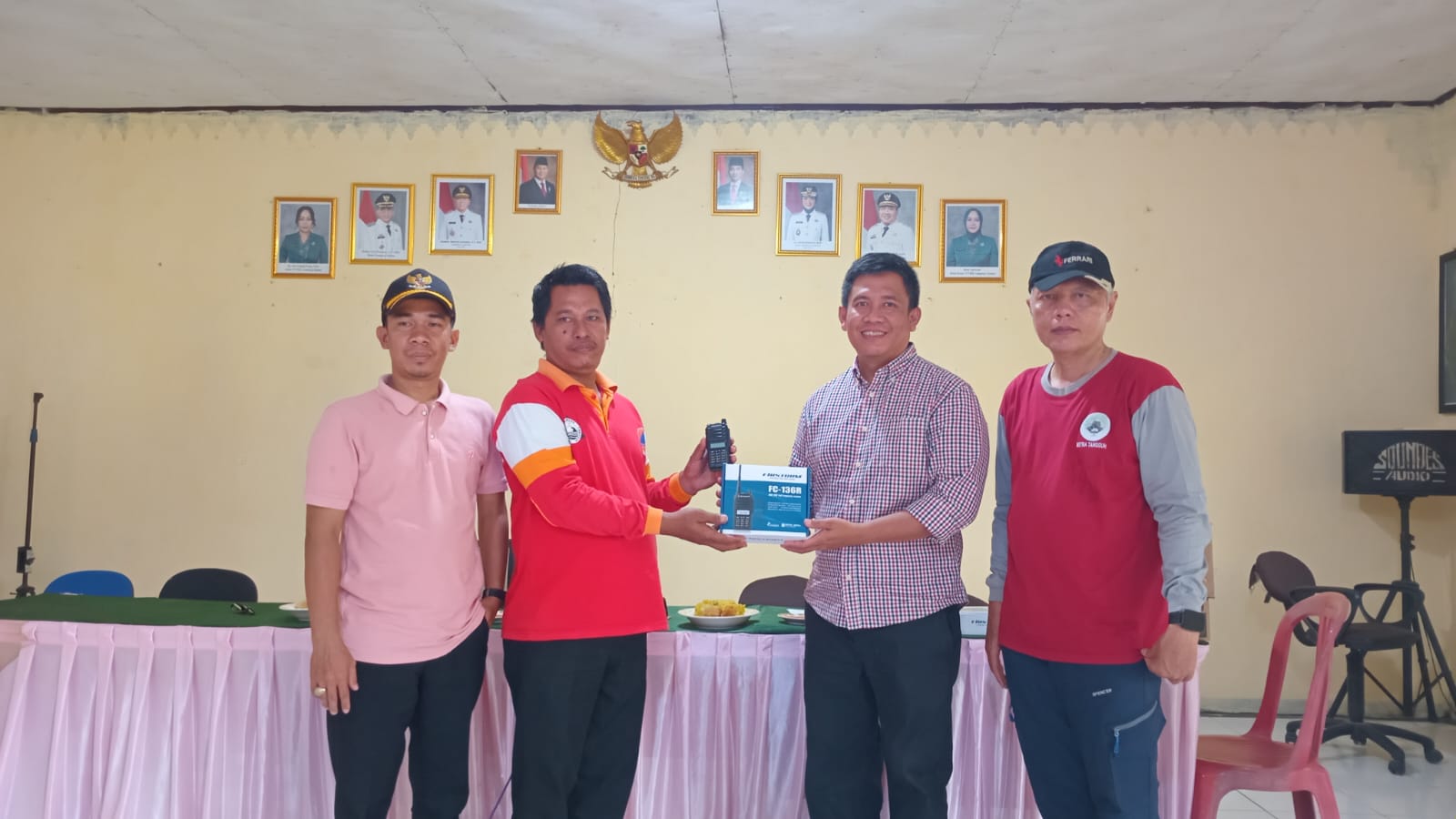 Empat pria berdiri di dalam sebuah ruangan pertemuan dengan latar dinding berwarna krem dan deretan foto pejabat serta lambang Garuda di bagian atas. Dua pria di tengah sedang menyerahkan dan menerima satu unit handy talky beserta kotaknya sebagai simbol serah terima. Di depan mereka terdapat meja panjang dengan taplak merah muda, sementara di sisi kanan terlihat kursi plastik dan perangkat pengeras suara.