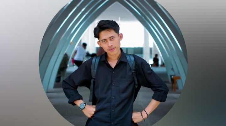 Seorang pria muda mengenakan kemeja hitam dan ransel berdiri dengan pose tangan di pinggang di bawah struktur lengkung modern, dengan latar belakang area terbuka dan beberapa orang di kejauhan.