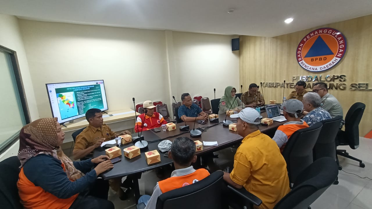 Foto suasana rapat di kantor BPBD Lampung Selatan. Belasan peserta duduk melingkari meja oval besar yang dilengkapi mikrofon dan kotak makanan. Di dinding belakang terdapat logo BPBD berukuran besar dan tulisan "PUSDALOPS KABUPATEN LAMPUNG SELATAN". Sebuah layar monitor di sisi kiri menampilkan presentasi bertajuk "Peta Wilayah Rawan Tsunami Lampung Selatan".