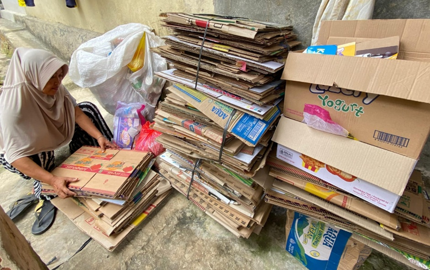 Aktivitas pemilahan sampah layak jual. Seorang ibu sedang merapikan tumpukan kardus kemasan (seperti kardus air mineral dan camilan) untuk disetorkan ke bank sampah atau pengepul. Di sebelahnya terdapat karung putih berisi botol plastik bekas.