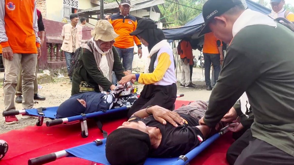 "Simulasi penanganan darurat medis di lapangan. Petugas dengan seragam oranye dan relawan sedang memberikan pertolongan pertama kepada dua orang yang terbaring di atas tandu lipat biru di atas karpet merah."
