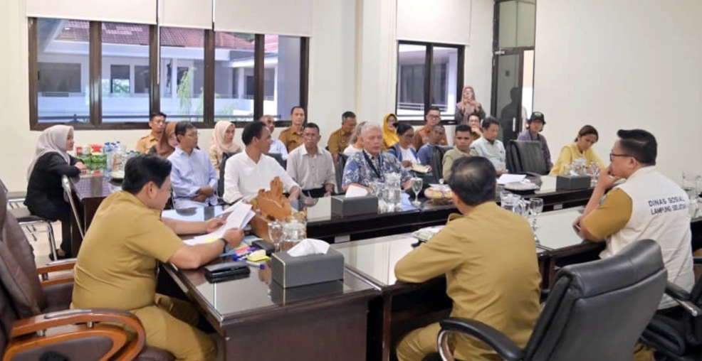 Sejumlah peserta rapat duduk mengelilingi meja konferensi di dalam ruang pertemuan kantor. Beberapa aparatur pemerintah daerah mengenakan seragam cokelat dan berdiskusi bersama peserta lain dalam suasana formal.